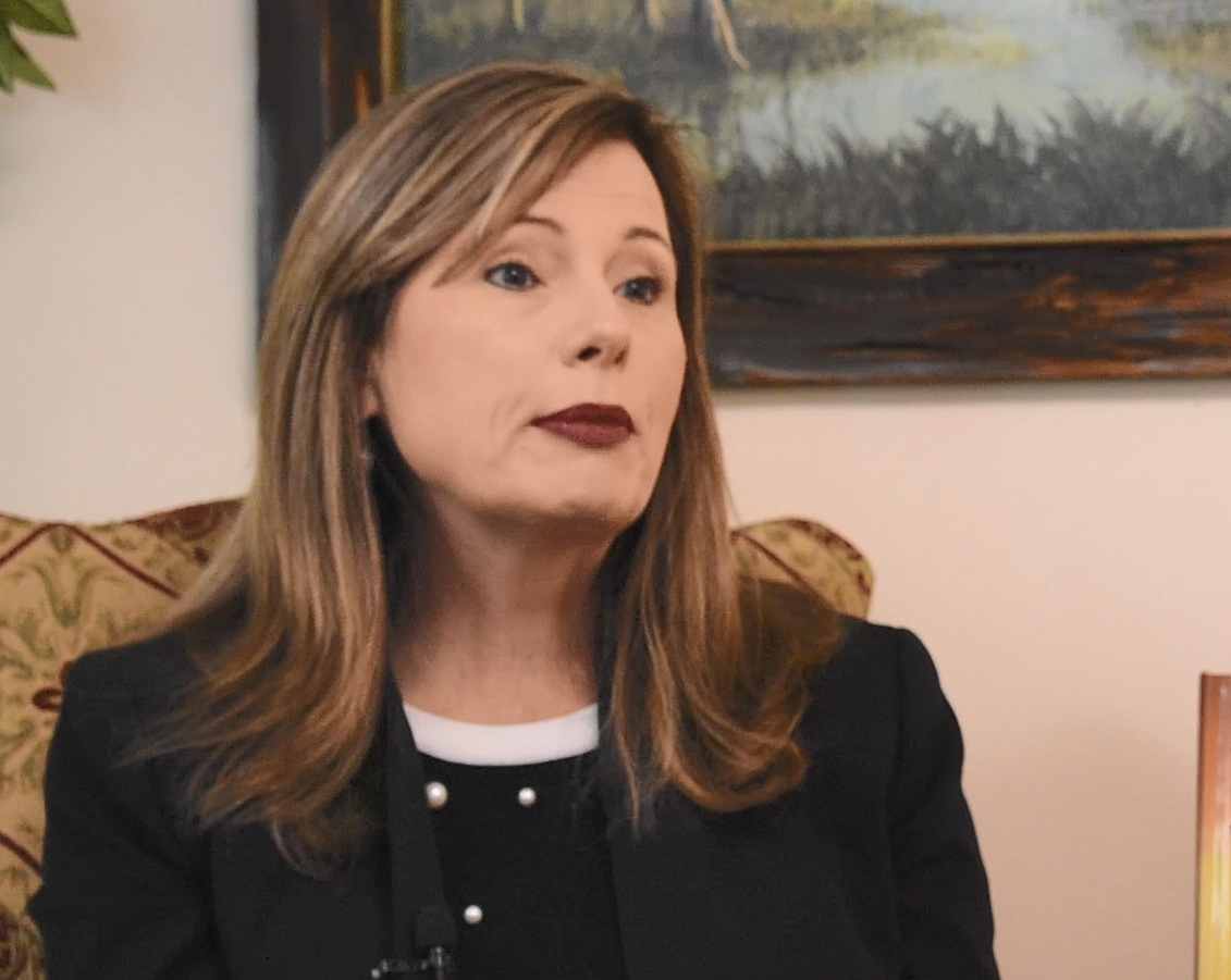 A woman with brown hair and black blazer sits in front of a landscape painting.