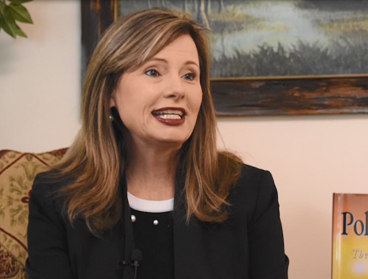 A woman with long brown hair and a black shirt speaks with an interviewer.