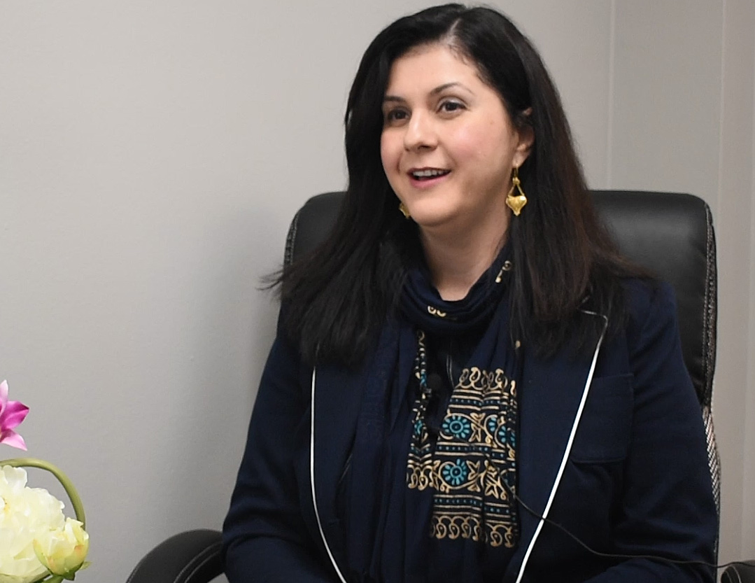 A woman with black hair sits in a leather chair beside some flowers.