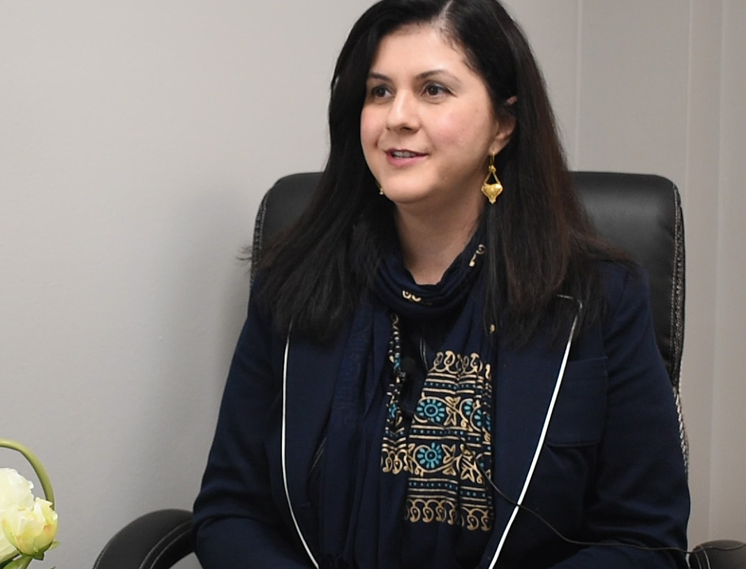 A woman with long black hair and a colorful scarf sits in a leather chair.