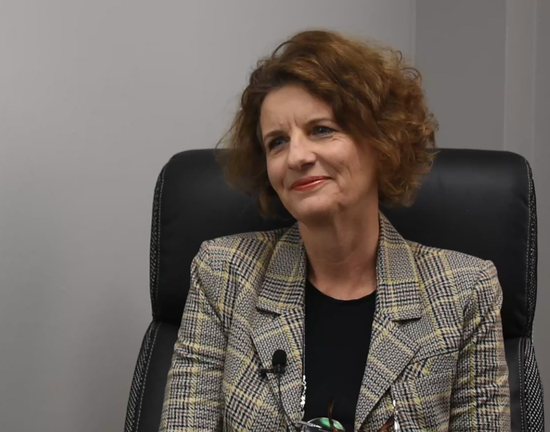 A woman with brown hair and a plaid blazer sits in a black leather chair.