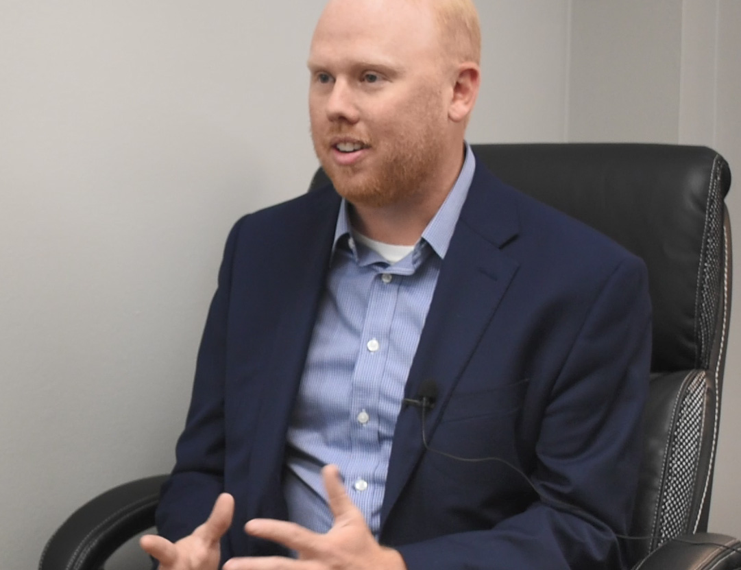 A man with red hair and blue blazer sits in a black leather chair.