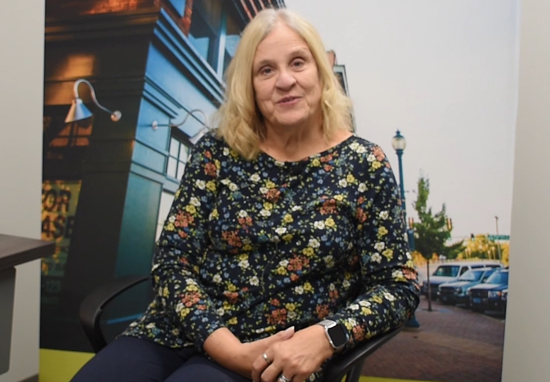 A woman with blonde hair and a black flowered shirt sits in front of a banner that shows a downtown main street.