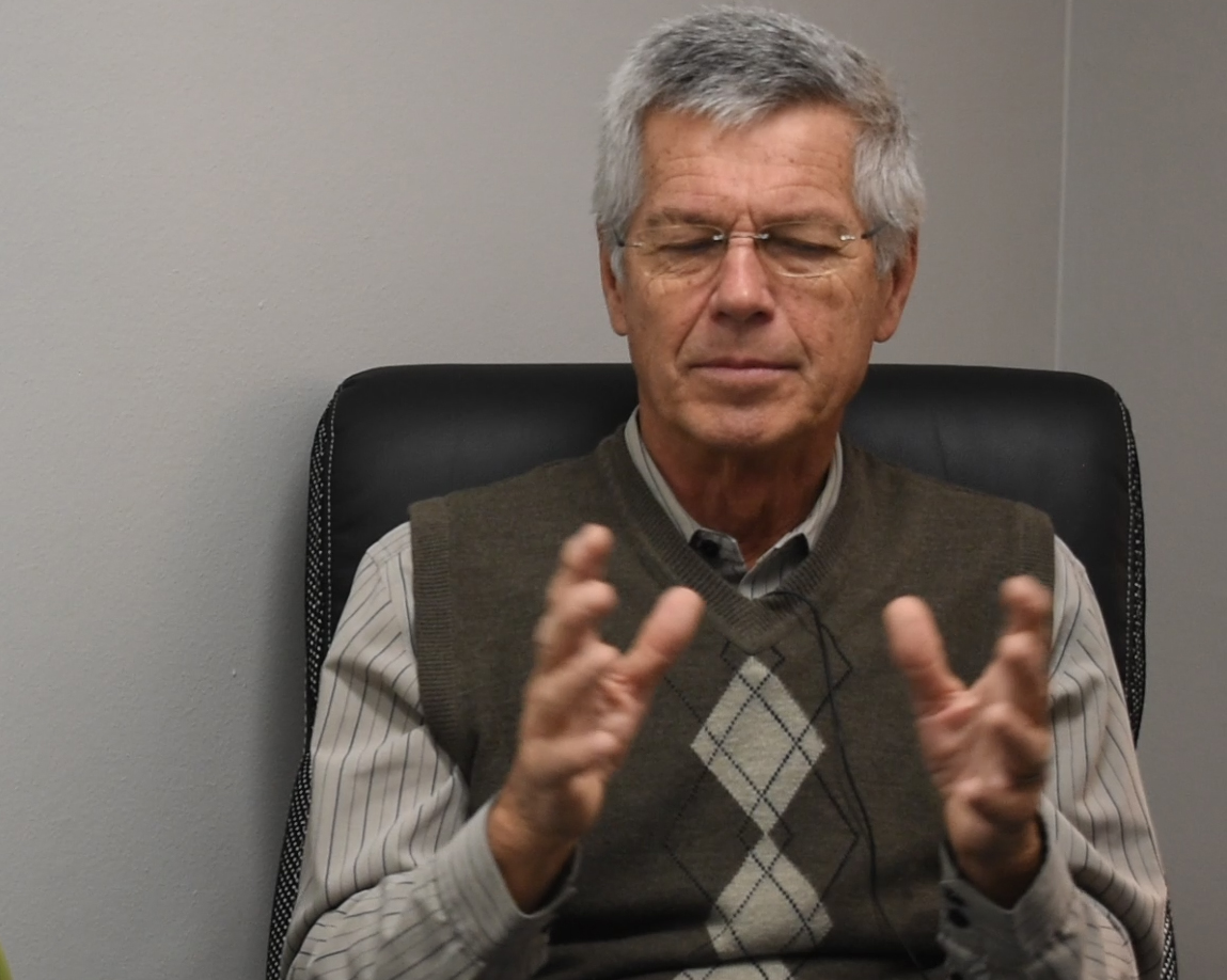 Jim has gray hair, glasses, and a brown sweater vest on.