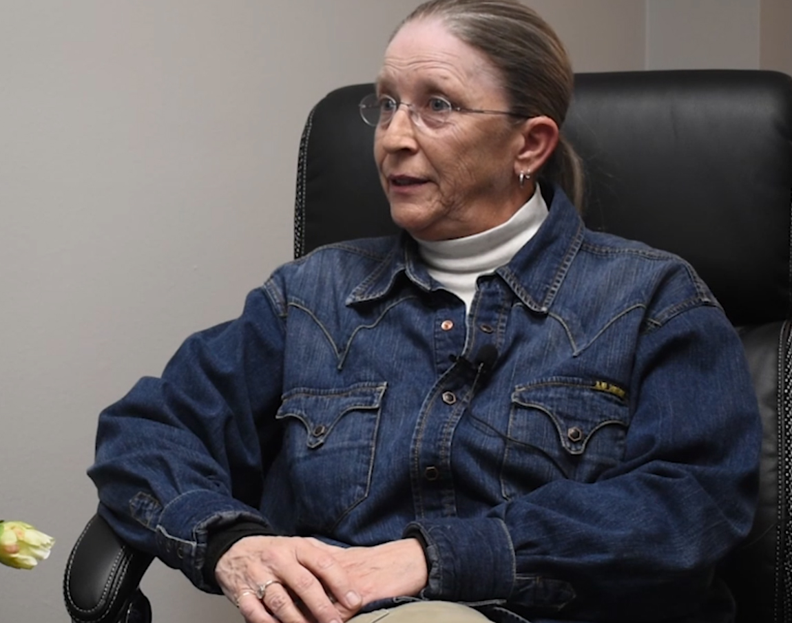 A woman with eyeglasses and her hair pulled tightly back wears a denim shirt.