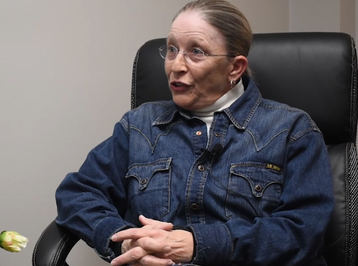 A woman eyeglasses and her hair pulled back tightly in a bun sits in a black, leather chair.