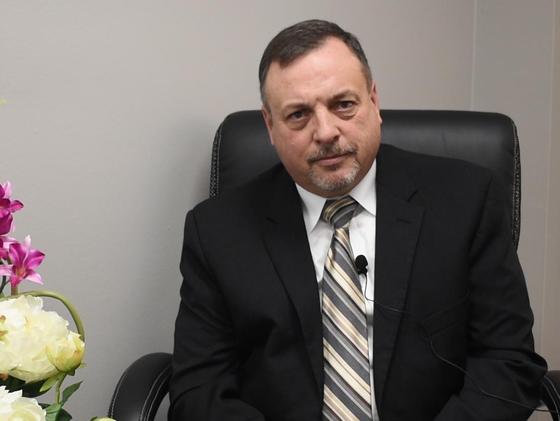 A man with short brown hair sits in a leather chair. He wears a suit and tie.