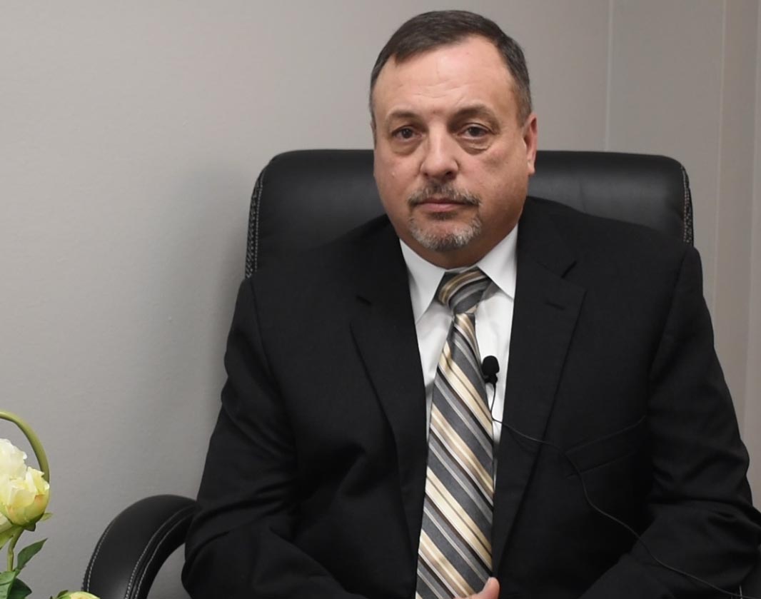 A man with brown hair and suit and tie on sits in a black leather chair.