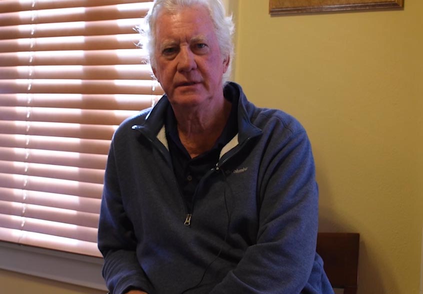 A man with a blue shirt and gray hair sits in a yellow room as he talks to an interviewer.