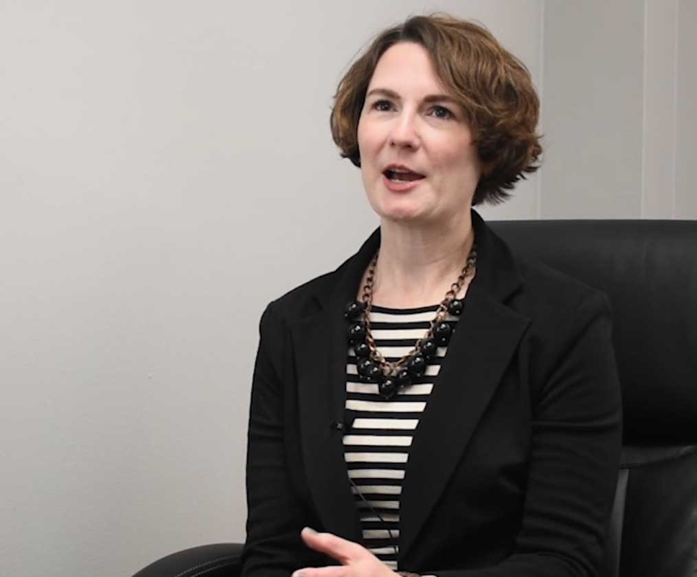 A woman with short brown hair, a black blazer, and striped black shirt sits in a black chair.