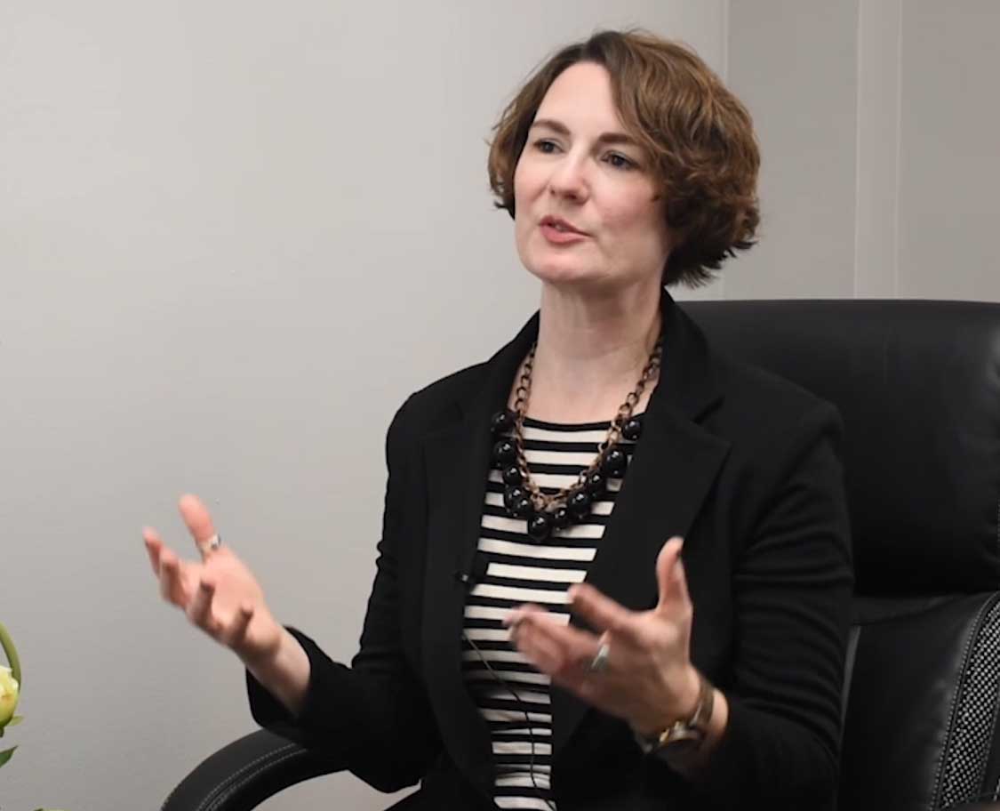 A woman with short brown hair and a black blazer sits in a black leather chair. 