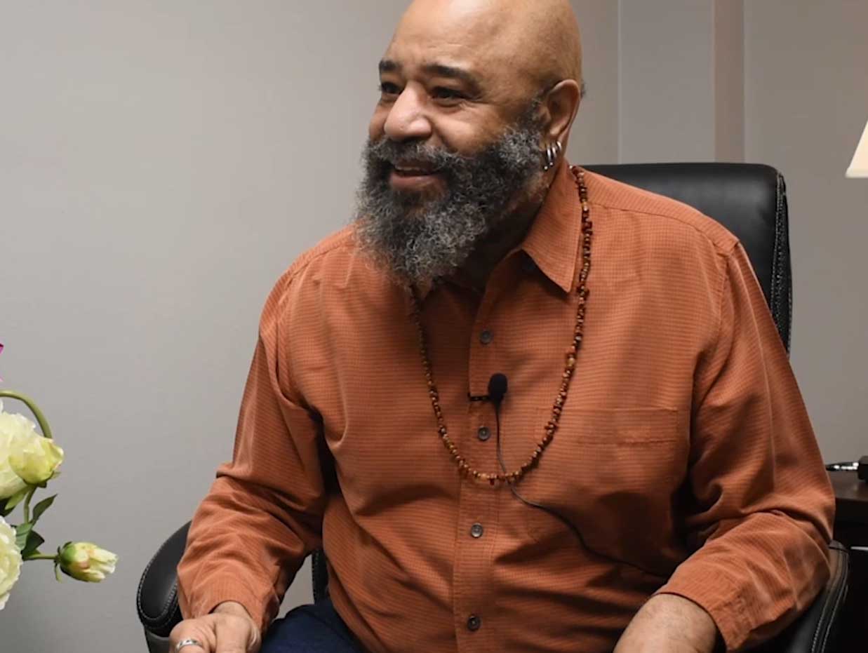 A man in rust colored shirt and a beaded necklace sits in a black leather chair.