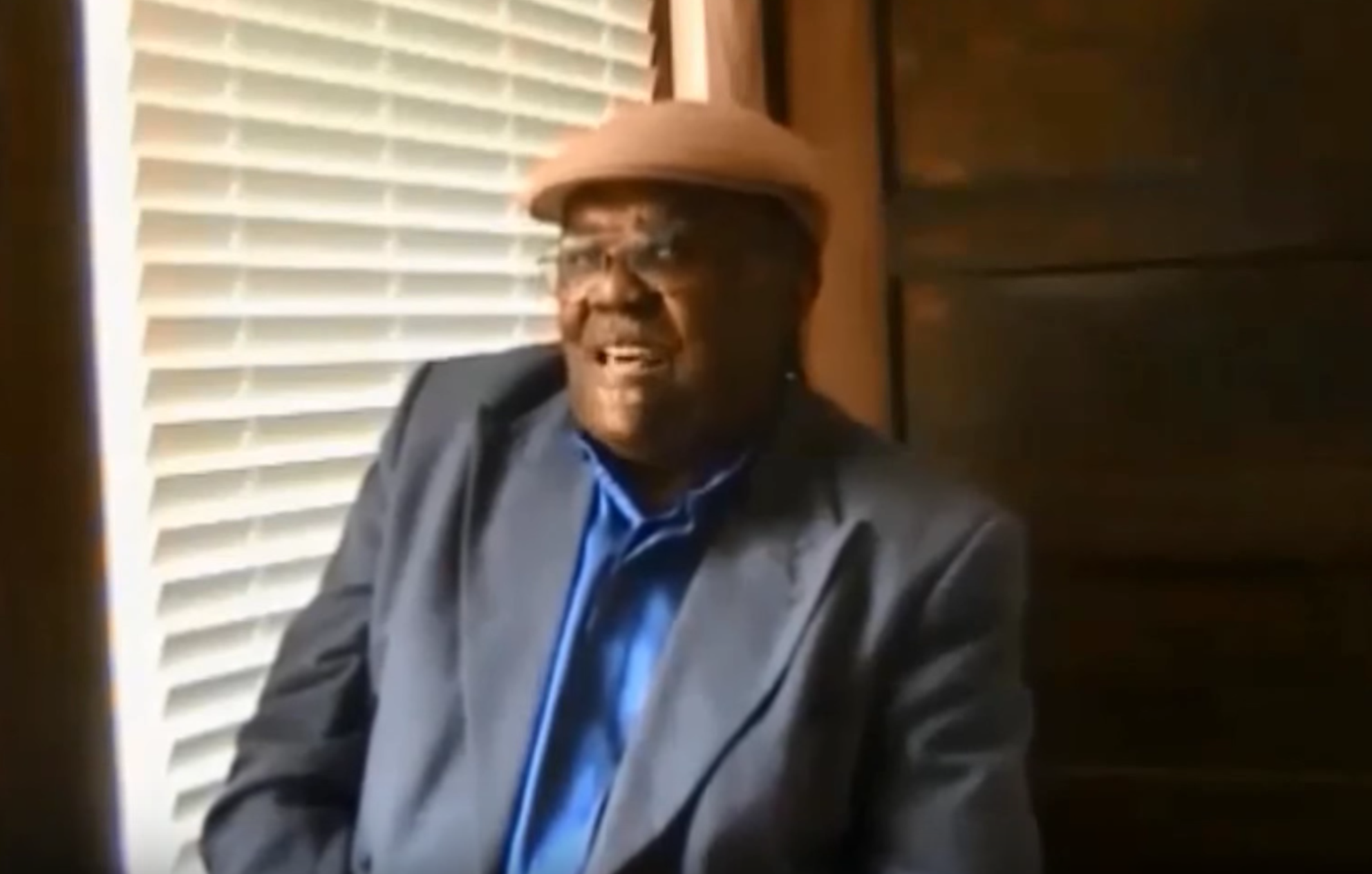 A man with a brown hat and a gray jacket sits in front of a window and talks to an interviewer