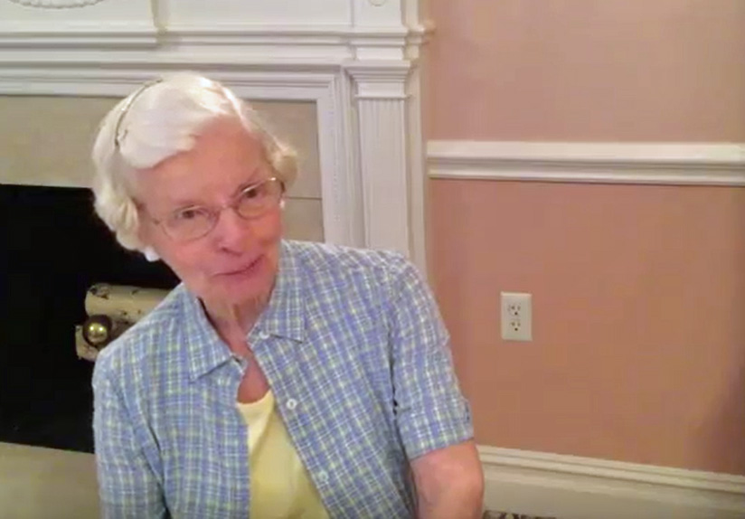 Miriam has short, white hair, a plaid blue shirt, and a yellow undershirt. She sits in a salmon-colored room with a fireplace.