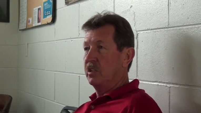 A man sits in front of a cinder block wall and chats with an interviewer.