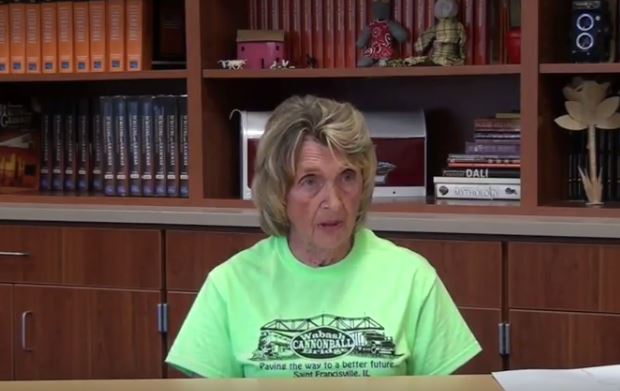 A woman with a bright green t-shirt sits in a library setting and is interviewed.