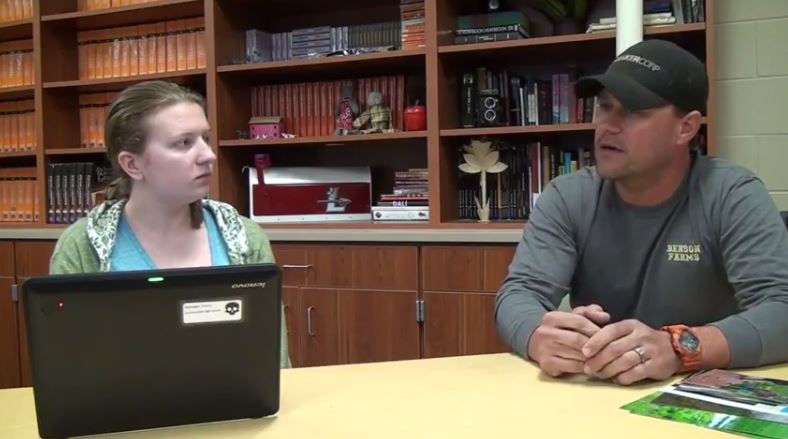 A young woman interviews a man with a ball cap in a library setting.