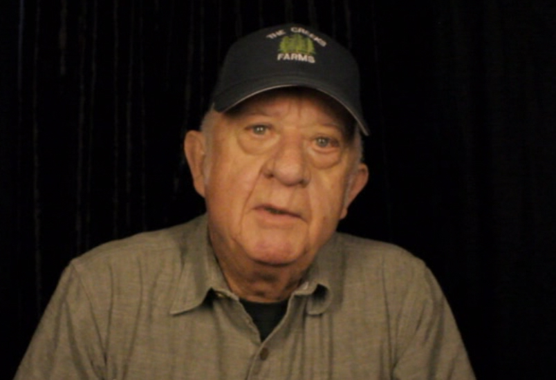 Don East of Alabama wears a black ball cap and a khaki colored button-down shirt against a black backdrop.