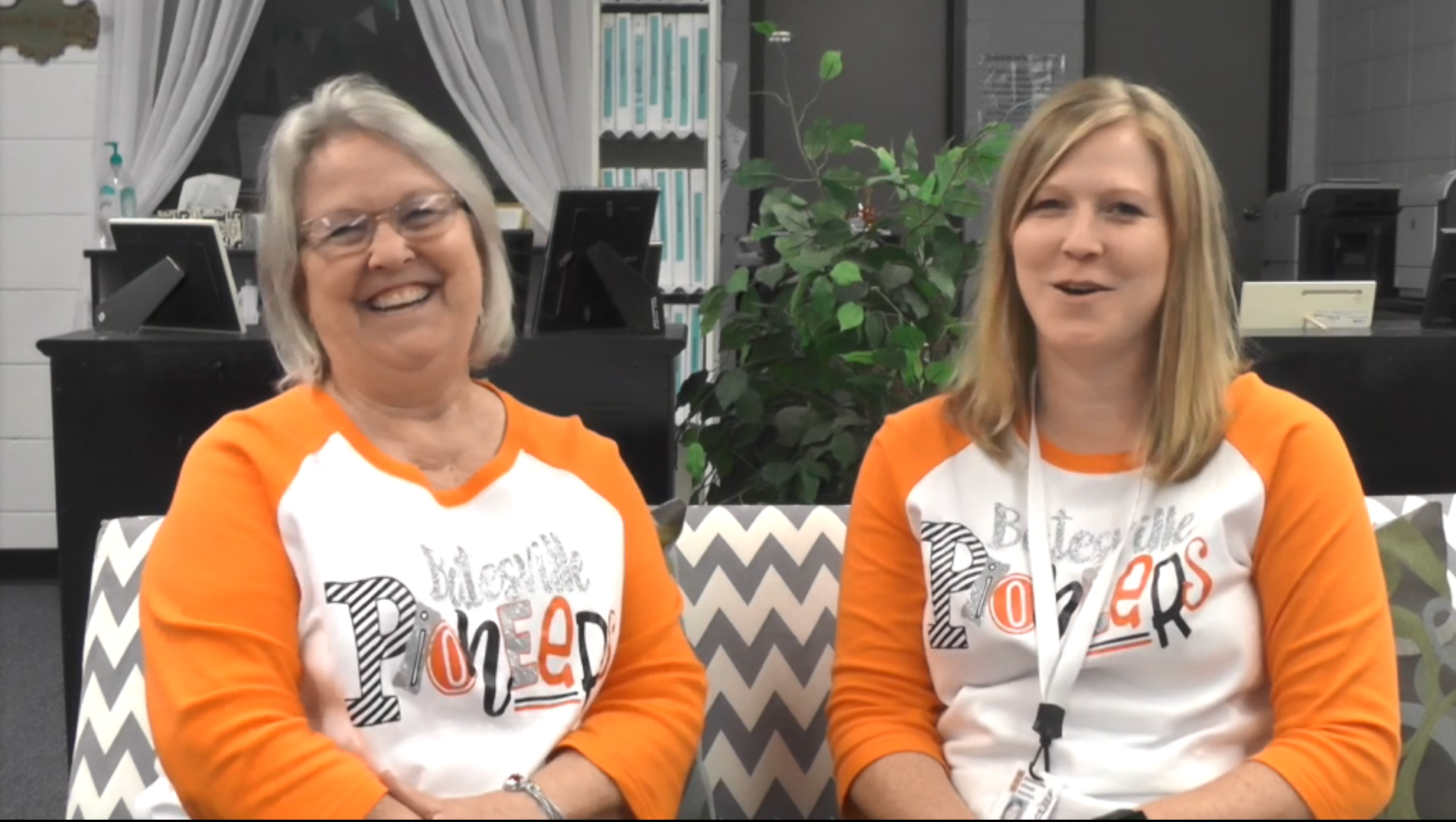 Two women, a mother and daughter, wear orange and white t-shirts and sit on a couch with gray, zigzag stripes.