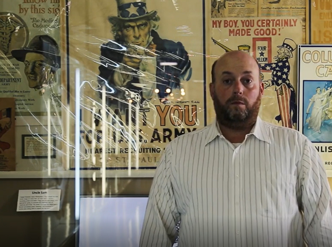 A man with a short-cropped beard stands in front of a museum case with war posters in it. 