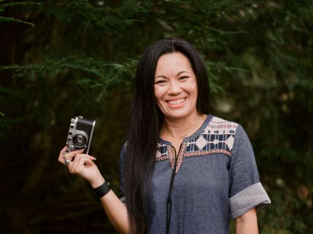 Jessica Maceda stands in a wooded area holding a camera.