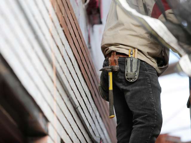A view from the waist down of a man with a toolbelt around his waist and working on siding on a house. Pexels stock photo.