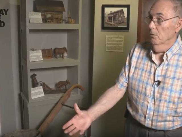 A man in a plaid shirt points to a plow and explains how it worked in the days before tractors.