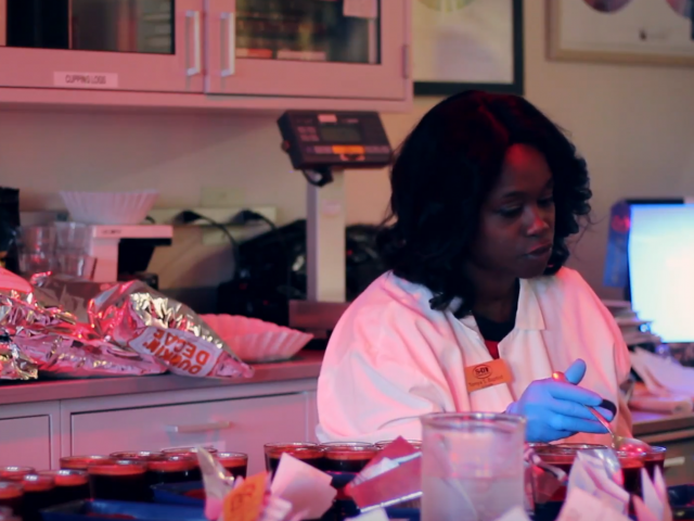 An African American woman with shoulder-length hair wears blue glues in a home kitchen as she stirs cups of coffee.