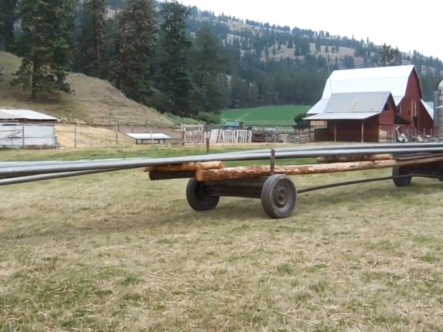 A flatbed attached to a tractor carries long pipes on the back of it as it pulls into a field on a farm.