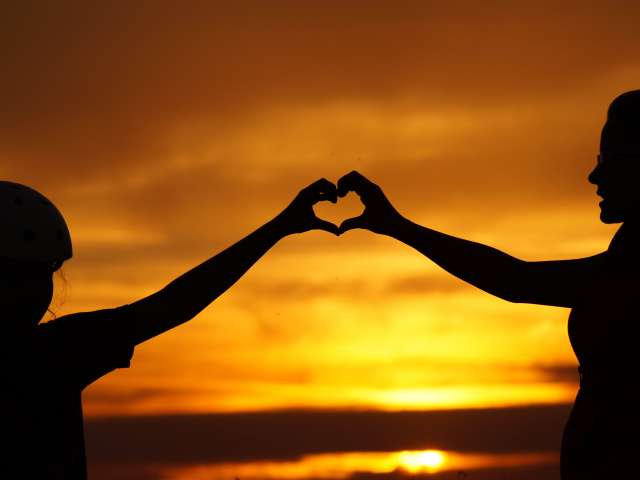 Two people, backlit by the setting sun, hold up their hands to form a heart. Pexels photo.