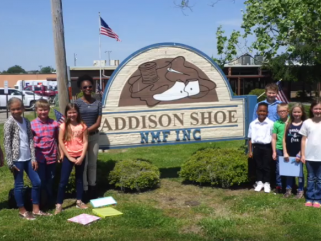 A group of students stand beside the Addison Shoe Company sign, outside the factory.