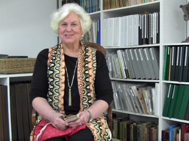 Nan has white hair, a black shirt, and a colorful scarf. She sits in a library.