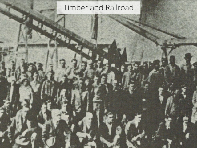 Black and white photo of a large group of men standing around machinery for timber.