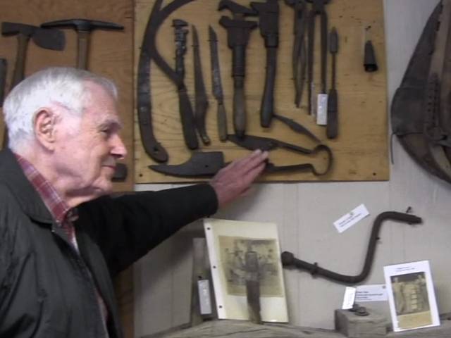 An elderly man stands next to a wall with many historic tools and points to a pair of large scissors.