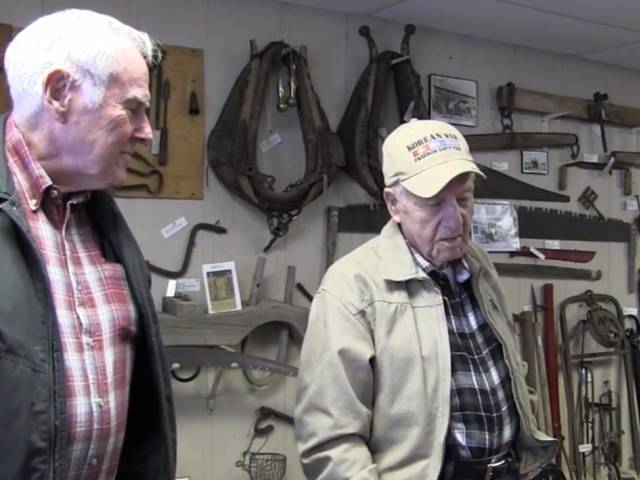 Two elderly men stand in a crowded museum space and talk about some of the objects on the walls, including old tools.