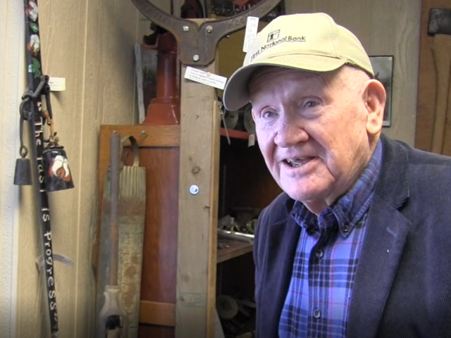 A man in a tan hat, plaid blue shirt, and blue blazer stands in front of a wall at a museum, hung with antique tools. 