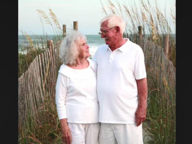 An elderly couple, wearing white, smiles at each other in a beachfront photo.