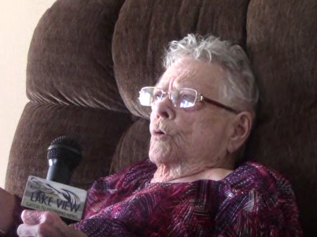 An elderly woman with gray hair and glasses sits in a plush brown chair and holds a microphone. 