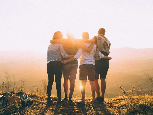 Four people with their backs facing the camera look out over a mountain at sunset. 
