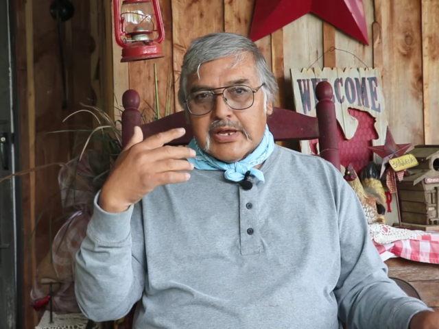 An man with gray hair, a gray shirt, and a turquoise bandana around his neck, sits on his porch and tells a story.