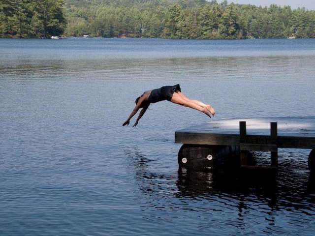 Wearing a black bathing suit, Mary dives off of a dock into a lake. 