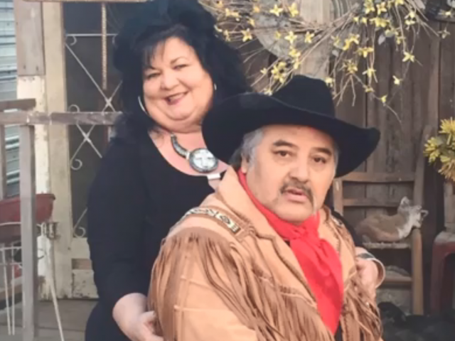A woman in a black dress stands behind a man in a Western hide coat and a black cowboy hat. 