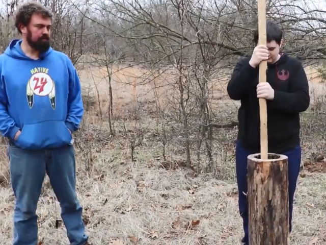 A man in a blue sweatshirt looks on as a young man uses a tall stick to mash something in a wooden log. 