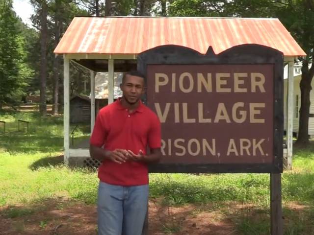 A young man in a red shirt stands in front of a park sign that reads, Pioneer Village, Rison, ARK.