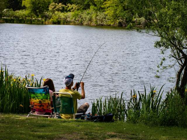 Two young people sit in lawn chairs by a lake and fish from the shore. 