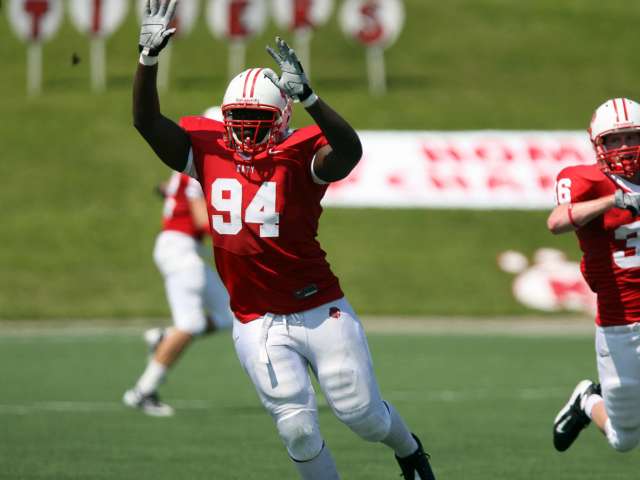 A young man, wearing a red football uniform, reaches up to catch a football on the field.