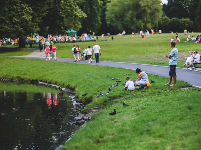 People walk on a paved park path while others sit by a pond on a summer day. 