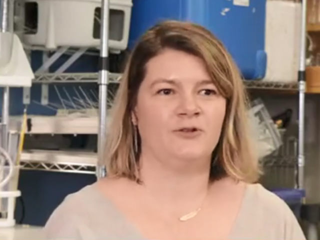 A woman with shoulder-length brown hair and gold necklace speaks in a lab.