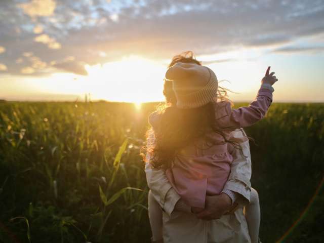 An adult carries a child on her back through a cornfield.