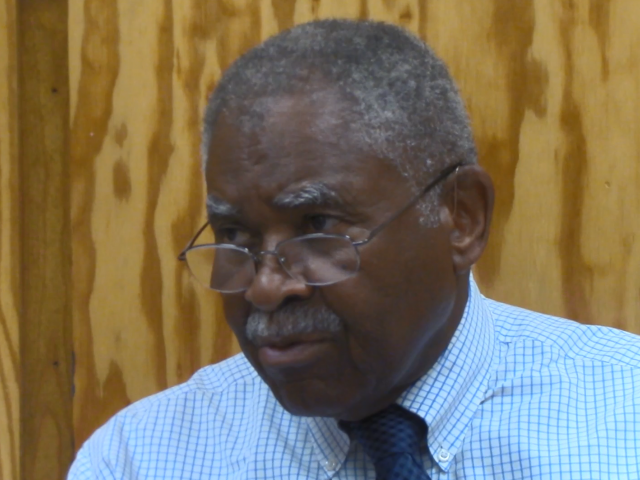 George Wilson wears glasses and a blue, striped dress shirt and blue tie. He sits in front of a wood-paneled wall. 